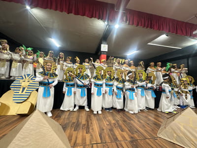 The image displays an indoor scene in Salé, Morocco, featuring approximately 25 to 30 children, primarily of elementary school age, dressed in ancient Egyptian-themed costumes. The children stand in rows on a raised wooden stage or platform, facing forward. Their attire consists of white tunics, gold and blue decorative collars, belts, and elaborate headpieces, including Nemes-style headdresses. Several children hold staff-like props. The backdrop behind them is dark, displaying the word "EGYPT" in stylized white lettering and a green sign with Arabic text. Overhead lighting is visible, and red pleated curtains adorn the upper background, indicating a performance venue. In the foreground, on a wooden floor, are two cardboard pyramid props and a large, flat cardboard cutout depicting a pharaoh's mask. The children appear to be posing for a photograph during a school play or cultural event.