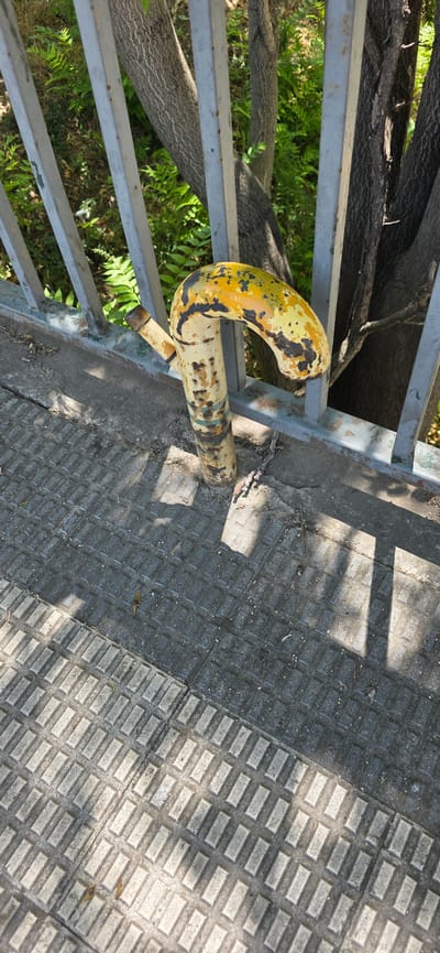 A close-up, high-angle shot features a bent, yellow and black painted metal pipe protruding from a concrete sidewalk. The pipe has a curved section, resembling a hook, and shows signs of rust and peeling paint. It is located next to a metal railing, beyond which is lush green vegetation and tree trunks. The sidewalk is paved with textured rectangular tiles. The scene is illuminated by sunlight, casting shadows across the ground. This image was captured in Huesca, Spain.