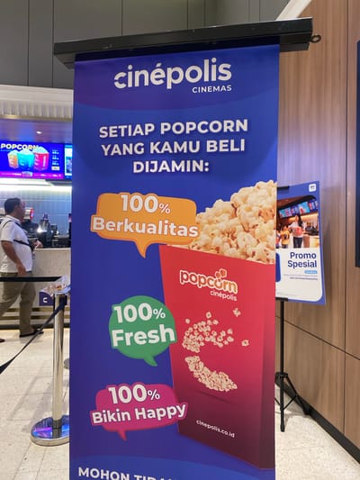 The image shows a vertical banner for Cinépolis cinemas in Banjarbaru, Indonesia, advertising popcorn. The banner is predominantly blue and features text in white and orange. The top of the banner displays the Cinépolis logo and the word "CINEMAS". Below this, the text "SETIAP POPCORN YANG KAMU BELI DIJAMIN:" is prominently displayed, meaning "Every popcorn you buy is guaranteed:".
The banner highlights three key features of the popcorn: "100% Berkualitas" (100% Quality) in an orange speech bubble, "100% Fresh" in a green speech bubble, and "100% Bikin Happy" (100% Makes Happy) in a pink speech bubble. An illustration of a red popcorn box filled with popcorn is on the right side of the banner, with a scattering of popcorn kernels below it. The website "cinepolis.co.id" is also visible at the bottom of the banner.
To the left of the main banner, a digital screen shows prices for popcorn and drinks, with options like "1 Drink" and different popcorn sizes. A man in a white shirt and khaki pants is standing near a counter, appearing to be a customer or employee. Metal stanchions with dark ropes are present, indicating a queueing area.
A second, smaller banner is partially visible to the right of the main banner. This banner has a white background and features text in blue and orange. It advertises "Promo Spesial" (Special Promo) and includes a QR code and a website address. The BRI (Bank Rakyat Indonesia) logo is visible at the top of this smaller banner, suggesting a partnership.
The overall environment suggests a cinema lobby or concession area, with tiled flooring and wooden-paneled walls in the background. The lighting is bright, typical of an indoor public space. The location is specified as Banjarbaru, Indonesia.
