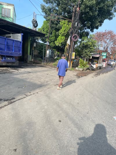The image shows a man walking down a street in Bandung, Indonesia, at the location -6.927, 107.584. He is wearing a blue shirt and brown pants. The street is paved with concrete and has a few small cracks. The man's shadow is cast in front of him. There is a truck parked on the left side of the street and some trees and vegetation on the right side. The scene is a mix of residential and commercial areas. The weather appears to be sunny and clear. There are several power lines and poles in the background. The man is walking toward the right of the image.
