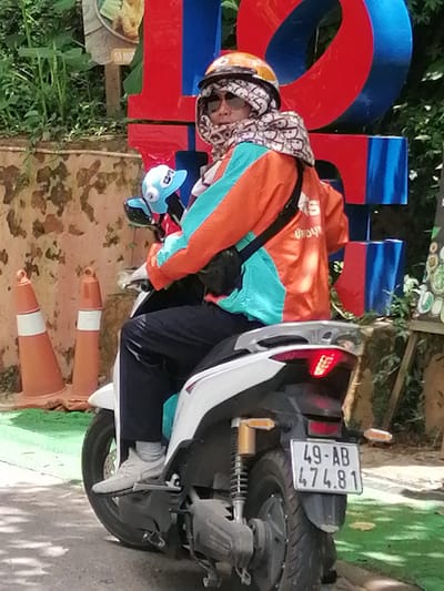 A person wearing a helmet, sunglasses, and a patterned scarf over their face is riding a white scooter in Da Lat, Vietnam. The rider is also wearing an orange and teal jacket and dark pants. The scooter has a license plate reading "49-AB 474.81". In the background, there are large red and blue block letters, two orange traffic cones, and lush green foliage. The ground appears to be paved with some areas of green artificial turf. The overall scene suggests a person on a scooter in an urban or tourist area.