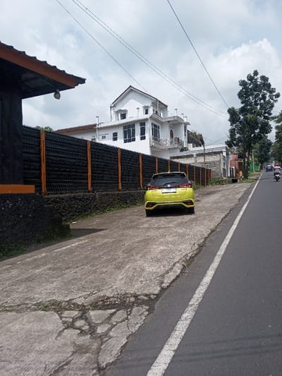 The scene depicts a daylight street view in Khajuripada, India, featuring a parked vehicle and surrounding urban structures. A bright yellow Toyota Yaris hatchback, bearing license plate "F 1242 YJ," is parked rear-facing on a concrete surface to the left of an asphalt road. The concrete surface exhibits cracks and wear. To the left of the parked car, a low, dark, textured wall supports a dark mesh fence with orange vertical posts. Behind this fence stands a prominent two-story white house with a red-tiled gabled roof, multiple windows (some tinted), a balcony, and white decorative trim. In the immediate foreground on the far left, a dark-colored structure with an orange-brown roof overhang and a mounted light fixture is visible. The asphalt road on the right features a solid white line marking its edge. Further down the road, traffic is visible, including small vehicles, possibly motorcycles or scooters, moving away from the viewer. A tall, leafy tree stands on the right side of the road in the mid-ground. The sky above is partly cloudy. No individuals are present in the immediate foreground.