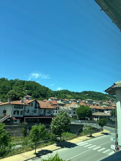 The image shows a panoramic view of Zlatograd, Bulgaria, on a clear, sunny day. The foreground features a paved street with a pedestrian crossing and sidewalks lined with yellow and grey tiles. A metal railing separates the sidewalk from a grassy area and a shallow, rocky stream.
In the midground, several buildings with red-tiled roofs are visible. One prominent building on the left has a sign that reads "КЪЩА ЗА ГОСТИ ТОНИ" (House for Guests Toni) and another smaller sign above it. This building also features an external staircase and several air conditioning units. Across the street, a bridge with an arched stone support spans the stream. A silver car is crossing the bridge.
The background is dominated by a lush green hillside covered in trees. The hillside is dotted with more houses, also featuring red-tiled roofs, as the terrain rises. The sky is a vibrant, cloudless blue with a few wispy white clouds forming a feathery pattern high up. A portion of a concrete structure with dark supports is visible in the top right corner of the image, suggesting a balcony or overhang. The overall scene depicts a picturesque town nestled within a natural, hilly environment.