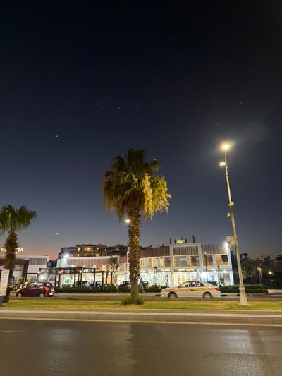 The image depicts a nighttime scene on a street in Sharm El Sheikh, Egypt. A tall street lamp illuminates the area with warm light. In the foreground, a palm tree with green fronds stands prominently. To the right, a modern building with multiple levels houses shops and businesses, indicated by illuminated windows and signage.
Visible text on the building includes "KAREM", "JAZZCO", "SUPER MARKET", "FIX PRICE", "RIHA", "ANTHEKA", and "RIHA". The storefronts suggest a commercial area. A white car with colorful decals is parked in front of one of the shops, and a red car is visible further down the street. The street itself appears wet, suggesting recent rain or mist. The sky is dark blue, with a few faint lights or stars visible. The overall ambiance is that of a lively commercial street at night, likely a tourist area given the location context. The lighting creates a somewhat atmospheric and inviting scene.