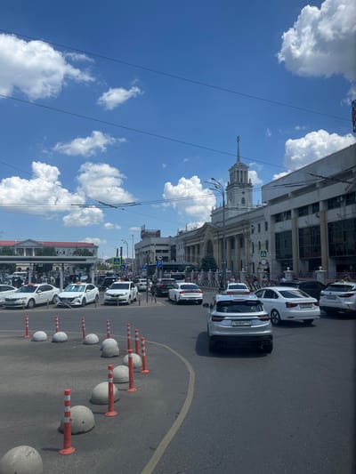 This media file depicts a street scene in Krasnodar, Russia, under a bright, cloudy sky, suggesting it's daytime. The perspective is from a slightly elevated, horizontal viewpoint, showing a line of white cars parked along the right side of the road, interspersed with concrete bollards and orange and white striped traffic cones. The cars are modern, with several appearing to be SUVs or sedans. The license plates of some cars are visible, with the format suggesting Russian registration. One plate reads "C468BP 193".

To the left of the road, a grand, multi-story building with classical architectural elements dominates the scene. It features columns, arched windows, and a prominent tower with a spire. The building appears to be a significant public structure, possibly a government building or a historical landmark. Power lines and streetlights are visible in front of the building.

Further down the street, more cars and buildings are visible, indicating a bustling urban environment. There are signs of pedestrian activity, with people seen walking on the sidewalk. The overall atmosphere is one of a typical day in a Russian city, with cars, architecture, and a clear sky. The weather appears to be pleasant, with fluffy white clouds scattered across a vibrant blue sky.