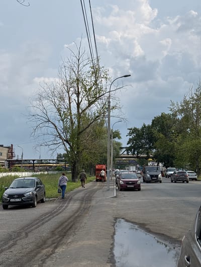 The image depicts a street scene in Izhevsk, Russia. In the foreground, a dark gray sedan is parked on the left side of a dirt road, with visible tire tracks on the surface. Two individuals are walking away from the camera on the dirt road towards the right. One is a woman wearing a gray hooded sweatshirt and blue jeans, carrying a black bag. The other is a man in a brown jacket and dark pants. Further down the road, a red truck with an orange bed is visible. The road transitions to asphalt and traffic can be seen, including several cars and a dark van. Trees line the right side of the asphalt road, and on the left, industrial pipes and a building with stairs are present. The sky is overcast with white and gray clouds.
