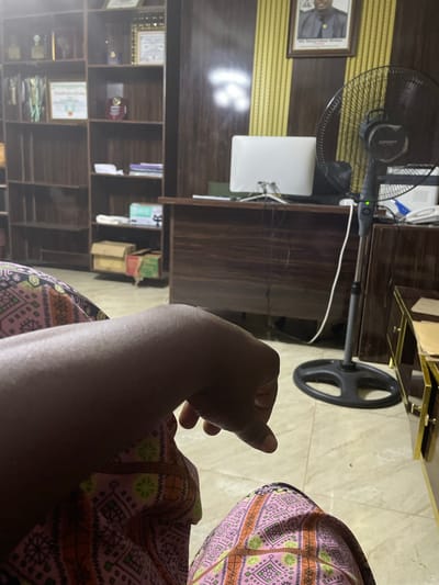 The image captures an indoor scene within what appears to be an office or study in Kwamba, Nigeria. The foreground features a close-up of a person's arm and leg, clad in vibrantly patterned pink and green fabric. The person's hand is visible, with fingers slightly curled and resting in a relaxed posture.
The background reveals a well-furnished room. To the left, a tall, dark wood bookshelf is filled with various items, including framed certificates and trophies, suggesting an academic or professional achievement display. Across from the bookshelf, a dark wood desk is visible, on top of which sits a white computer monitor and a telephone. A black oscillating fan stands to the right of the desk, its green indicator light illuminated. Behind the desk, the wall is decorated with yellow striped wallpaper above a section of dark wood paneling. A framed portrait of a man in formal attire hangs prominently on the upper part of the wall. Adjacent to the desk, a piece of furniture with a dark wood top and gold-trimmed drawers is partially visible. The floor is tiled with a light-colored, patterned material. Several cardboard boxes and other items are stacked on shelves in the far background.