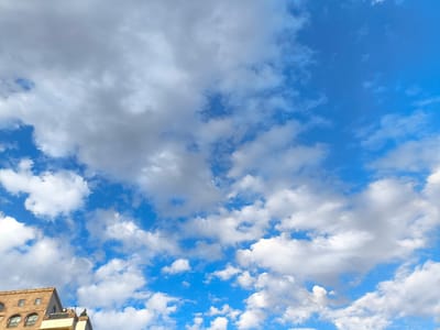The image shows a bright blue sky with scattered white and gray clouds. In the lower left corner, the top of a building with arched windows and a tiled roof is visible. This building appears to be in a city. The architectural style suggests it could be in Yerevan, Armenia, known for its distinctive stone buildings and historical architecture. The overall scene depicts a daytime sky with some cloud cover, likely a pleasant weather day.