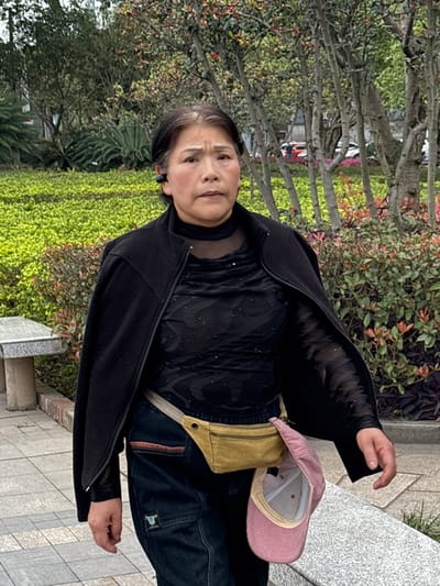 An East Asian female, appearing middle-aged or older, is depicted walking on a paved pathway in a public green space in Jiangjin, China. She is oriented slightly to the viewer's right, looking forward with a neutral expression. Her dark hair exhibits some grey at the temples. A black earbud is positioned in her left ear. She wears a black, open-front jacket draped over her shoulders, revealing a black, sheer, long-sleeved high-neck top beneath, which features subtle reflective details. Her lower attire consists of dark blue denim jeans. A khaki-colored fanny pack is secured at her waist, with a light pink corduroy baseball cap attached and hanging inverted on her right side. To the left of the pathway, a grey stone bench is partially visible. The background is composed of manicured green shrubs and bushes, punctuated by several slender trees displaying sparse foliage with some reddish coloration. Distant background elements include faint urban structures and a partially visible red vehicle. The scene is illuminated by natural, ambient light.
