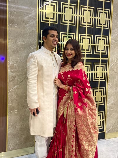In Dhaka, Bangladesh, a man and a woman pose for a photograph indoors. The man lies horizontally, wearing a light beige sherwani over white pants, holding a black object in his right hand. The woman lies atop him, draped in a red and gold saree. She has bangles on her right wrist and henna on her hand. Both are smiling and looking towards the camera. Behind them is a decorative wall panel with a black and gold geometric design. The floor is covered with light brown marble tiles with brown trim. A blue light is visible at the bottom left of the frame.