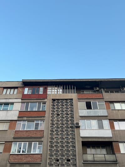 The image shows a section of a Brutalist-style apartment building in Pazardzhik, Bulgaria, set against a clear blue sky. The building's facade is characterized by its exposed concrete, alternating with brickwork and numerous windows. A prominent feature is a large, decorative concrete panel with a repeating geometric pattern running vertically along the center of the visible section. Several balconies are visible, some with enclosed windows and others with railings. Air conditioning units are mounted on the exterior walls. The architectural style suggests it is likely a Soviet-era or post-Soviet era building.