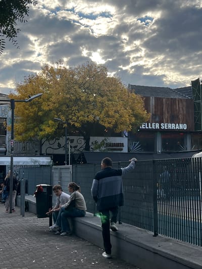 The image is a street scene in Buenos Aires, Argentina, on a cloudy day. A building is labeled "KELLER SERRANO." A large tree with yellow leaves stands near the building. Several people are present. One man is standing near a metal fence, with his arm raised. Two people, one wearing a white shirt and the other a light sweater, are positioned near a trash can on the sidewalk. Further down the street, there are additional people visible. The ground is paved with bricks.