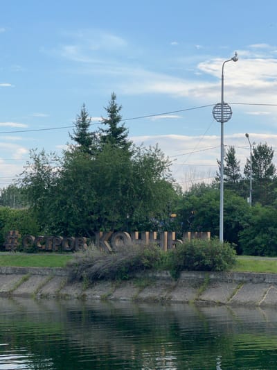 The image shows a body of water in the foreground, with a concrete embankment behind it. On the embankment, there is lush green vegetation and large, three-dimensional wooden letters forming the word "ОСТРОВ КОЛ" which translates to "ISLAND KOL". Behind the letters, several tall evergreen trees are visible, along with a streetlamp with a globe-like fixture and a bird perched on top. The sky is partly cloudy and blue. This scene is likely located in the city of Irkutsk, Russia, possibly near the Angara River or one of its tributaries, given the presence of such a prominent water feature and urban elements.