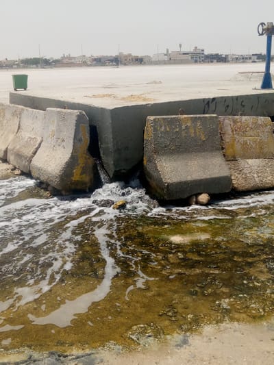 The image shows a discharge of water flowing under a concrete barrier into a shallow body of water. The barrier is composed of interlocking concrete blocks, some marked with yellow paint, and appears weathered. The water is foamy and flows over a bottom with visible sediment and possible algae growth. In the background is a flat, paved area with a green trash bin and a blue utility structure. Beyond this area, a cityscape of low-rise buildings can be seen, suggestive of an urban or suburban environment. Based on the prompt information, the location is Al `Aziziyah, Saudi Arabia. No people are visible in the image.