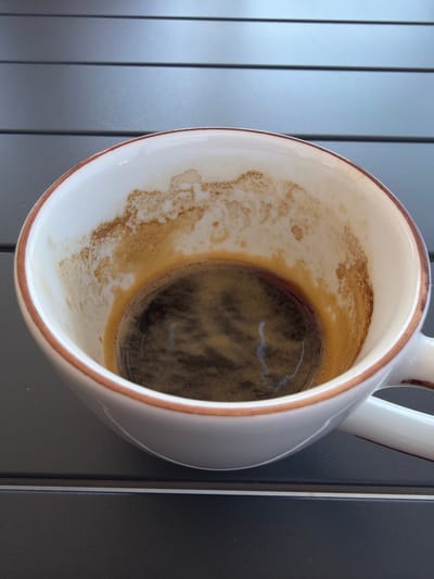A close-up, overhead shot shows the inside of a white ceramic coffee cup with a thin brown rim. The cup contains a small amount of dark liquid, likely espresso, and the sides are stained with light brown coffee residue. The cup rests on a dark, slatted surface, possibly a table or deck. The background is out of focus, showing more of the slatted surface. There are no people or other objects visible. The location is the Sithonia Municipal Unit, Greece.