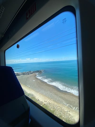 The image captures a view through a large, curved window, likely from a train, looking out at a beach and the ocean. The window is tinted blue, creating a serene atmosphere. The scene outside is a picturesque coastal landscape on a clear day. The ocean is a vibrant blue, with gentle waves breaking on a pebble beach. The beach is lined with stones and some patches of green grass. A rocky breakwater extends into the water, disrupting the smooth line of the shore. The sky is a brilliant, cloudless blue.
Inside the train, the dark interior frames the bright exterior view. The edge of a blue seat is visible in the lower right corner, suggesting the viewpoint is from a passenger. On the window itself, faint horizontal lines are visible, possibly heating elements or structural lines. There is also a circular red and black object adhered to the inside of the window, and a faint white circular sticker with text is visible near the top left of the window. The text on the sticker reads "SIEMENS Aqua-Frost 2012-02", indicating the manufacturer and potentially the year the window was made. On the interior paneling below the window, a red square sticker with a white arrow pointing upwards is visible, which could be an emergency exit or information sign. The overall impression is one of peaceful travel, with the stunning coastal scenery offering a sense of calm and relaxation. The time of day appears to be daytime, given the bright sunlight and clear blue sky. The location is identified as Khostinskiy, Russia.