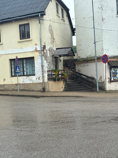 The image captures a street view in the city of Sabile, Latvia, focusing on the exterior of two adjacent buildings. The building on the left is painted a pale yellow and shows signs of wear, with peeling plaster revealing brickwork beneath. It features several windows with dark frames. A blue and white pedestrian crossing sign is visible on the left side of the image, in front of a window. Further to the right, a separate entrance with a small awning is visible, built against a stone wall and accessed by a set of stairs with a wooden railing. The building on the right is painted white and also exhibits peeling paint. A red and blue circular traffic sign, indicating no parking, is mounted on a pole in front of this building. The ground in the foreground is a wet asphalt road, reflecting the overcast sky. The overall scene depicts a somewhat aged urban environment with visible signs of weathering on the structures.