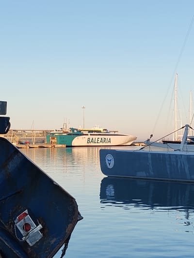 The image shows a harbor scene in Dénia, Spain, at dusk or dawn, evidenced by the soft, diffused light. In the background, a large, white passenger ferry with the name "BALEARIA" prominently displayed is docked at a pier. The ferry features green accents and a modern design. Midground shows one yacht and the masts of others and the foreground presents sections of a two smaller boats. The water is still and reflects the colors of the sky and the surrounding boats, contributing to the overall calm atmosphere of the harbor. The scene lacks people.