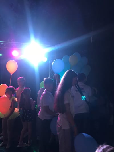 The image depicts a dimly lit indoor scene, likely a party or event, with several individuals, predominantly children, gathered. Prominent bright stage lights, emitting a strong blue and white glow, illuminate the scene from the upper left. In the foreground and midground, there are several colorful balloons, including orange, yellow, and blue, adding to the festive atmosphere.

The individuals present appear to be a mix of children and possibly some adults. Some children are seen in the foreground, facing the camera or in profile, with varying hairstyles and clothing. One child on the right is wearing a white polo shirt. Another child, further back and to the left, is wearing a yellow tutu-like skirt. A young girl with pigtails is visible in the midground, wearing a dark skirt with white polka dots. A young woman with long dark hair is in the center, facing away from the camera, with a white t-shirt. Another person, likely an adult male, is partially visible in the background, wearing a white t-shirt with text and a glowing circular light on the back.

The environment is dark, with the bright lights creating lens flares and casting shadows. The presence of balloons and the general ambiance suggest a celebration or entertainment event.

This image was taken at the "Morski Zvukove" (Sea Sounds) festival at the Sea Garden in Varna, Bulgaria, on August 12, 2017. The festival is an annual event held in the open-air theatre in Varna, featuring music and entertainment.