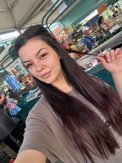 A young woman with long, dark hair and freckles takes a selfie in an outdoor market. She is wearing a light brown t-shirt. In the background, there are market stalls with various goods, including produce and bags. Several other people are visible, some browsing, some working at the stalls. Signs with text are also present. The overall setting suggests a lively market environment. The location is the "Tsar Simeon's Garden" market in Sliven, Bulgaria, specifically identified by the Cyrillic text on the sign "ОРАНЖЕ КРЪСТЕВИ" which translates to "Orange Tree Christian" and is a known vendor at this market.