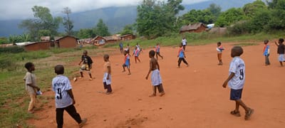 Children are playing soccer on a dirt field in Ngalula, Democratic Republic of the Congo. Approximately 15 boys are visible, some in soccer jerseys, others in casual clothing. In the background are small brick houses with thatched roofs, surrounded by green vegetation and mountains obscured by clouds. One adult male is standing near the houses. The ground is red clay, typical of the region. The sky is overcast.