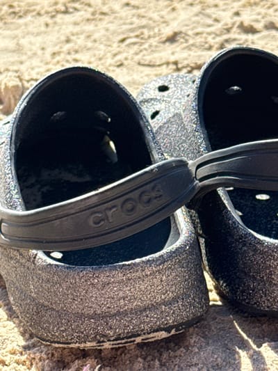 A close-up, overhead shot captures two glitter-covered black Crocs resting on a sandy surface, likely a beach in Tel Aviv, Israel. The Crocs are positioned side-by-side, with their strap closures visible in the foreground. The sand is light brown and appears textured, possibly from footprints or wind patterns. Sunlight casts shadows, highlighting the grainy texture of both the sand and the shoes. The left Croc is more prominently displayed, showing its interior and the embossed "CROCS" logo on its strap. The overall scene suggests a casual, outdoor setting.