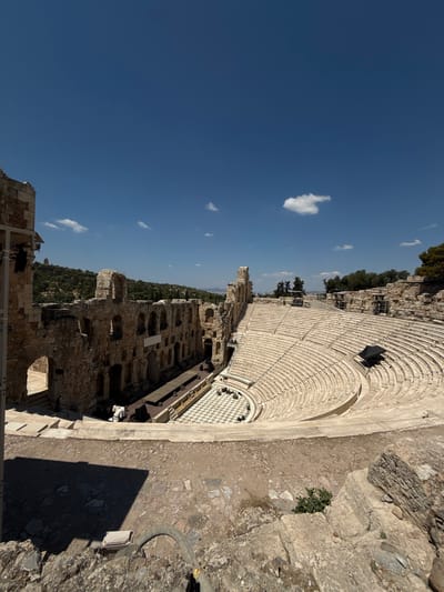 The image depicts the Odeon of Herodes Atticus, an ancient stone amphitheater in Athens, Greece, situated on the southwest slope of the Acropolis. The amphitheater's tiered seating, constructed from pale stone, curves around a semi-circular stage area. The stage itself is adorned with a checkered floor pattern. In the background, the ruins of stone structures, likely part of the ancient theater complex, stand against a backdrop of lush green hills under a clear blue sky with scattered white clouds. The foreground is dominated by rough, uneven terrain with large stones and a shadow cast from an unseen object. Two individuals are visible on the stage area, one standing and one seated, though their specific actions are not discernible. The overall scene suggests a historical landmark on a bright, sunny day.