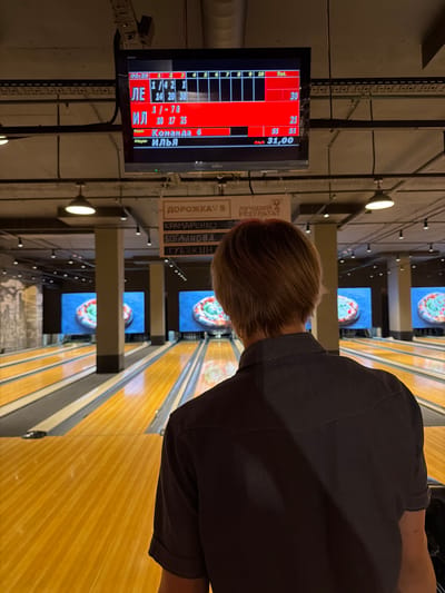 The image depicts a bowling alley, likely in Izhevsk, Russia, based on the Cyrillic text visible. In the foreground, the back of a person with light brown hair is shown, wearing a dark grey collared shirt. They are facing a bowling lane and looking towards a large screen displaying game scores. The screen shows a bowling score card with names and scores in Cyrillic, including "ЛЕ", "ИЛ", "Команда 6", and "Player ИЛЬЯ". The score card indicates frames 1 through 10 and totals. Further down the lane, two large screens are visible, each displaying a pizza on a blue background. The bowling lanes are illuminated and appear to be made of polished wood. Above the lanes, overhead lighting fixtures are visible, along with a visible section of the building's industrial ceiling structure. A sign with Cyrillic text, reading "ДОРОЖКА 5", is mounted on the wall above another display.