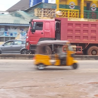 The image depicts a street scene in Dadi Riba, Nigeria. A red truck with a burgundy cargo container is prominent in the center of the frame. To the left of the truck, there are structures, including a building with a sign partially visible. The street surface is visible and appears to be textured. A gray car is seen parked beside the truck. A blurry yellow object is moving along the street next to the truck. The overall scene is somewhat blurred, indicating motion or a low-quality image.
