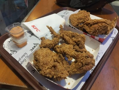 A tray of KFC fried chicken rests on a wooden table. The tray is lined with white paper featuring the KFC logo. In the foreground, a stack of three small, clear plastic containers holds dipping sauces. To the right of the sauces, a white KFC napkin is folded. Arranged in two white paper containers are several pieces of crispy fried chicken. The lighting suggests an indoor setting, likely a restaurant or food court. The overall scene depicts a meal ready to be consumed. No individuals are visible in the image.