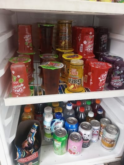 A refrigerated display showcases a variety of beverages. The top shelf is stocked with cups of Okky Maxx in assorted fruit flavors and G-Bean juice. Below, there are bottles of Pucuk Harum and Milk Jus, alongside various canned drinks, including soda and Nestle products. A container holds small packages of "Sudah Matang Tinggal Makan" snacks. The arrangement appears to be for retail sale, with the items neatly organized for customer selection.