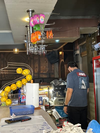 A man wearing a dark blue t-shirt with a "JAWS" graphic on the back and black pants stands with his back to the camera in what appears to be a small cafe or bar. He is standing near an espresso machine and to the right of a display of lemons in a metal rack. Behind the lemons is a roll of paper towels. To the far right of the frame, a red Coca-Cola refrigerator is visible. The background shows a wall with a decorative wallpaper of bookshelves and a hanging lamp. Several glasses are suspended from a rack overhead, along with brightly colored packets of what appear to be flavorings or sweeteners. The overall environment suggests a casual, perhaps slightly cluttered, establishment.