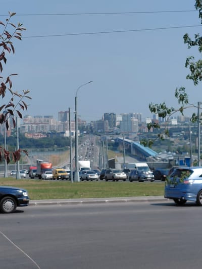 The image shows a panoramic view of a city skyline under a clear blue sky, likely taken from a highway or elevated road. In the foreground, on the right side, a blue SUV is parked or driving. The vehicle appears to be decorated with green foliage around its rear. The SUV is positioned on a paved road, with a grassy embankment behind it.
Further back, a traffic scene unfolds on a multi-lane road, possibly a bridge or elevated highway. Various vehicles, including cars and trucks, are visible. Some vehicles appear to be stopped or moving slowly, indicating traffic congestion. A yellow van is prominent among the vehicles.
In the middle ground, beyond the road, is a body of water, possibly a river, with more urban development on the opposite bank. This is followed by a dense cluster of buildings, suggesting a city center. The architecture of the buildings varies, with some appearing modern and others older. Green trees and vegetation are scattered throughout the cityscape, especially on the hillsides behind the main urban area.
In the background, the skyline is dominated by numerous buildings of different heights and styles. Power lines are visible in the sky. The overall impression is a bustling urban environment with significant infrastructure.
While the image doesn't contain explicit markers to pinpoint Novosibirsk, Russia, the architectural style of the buildings and the general landscape are consistent with many Russian cities, and this type of urban and infrastructure development is common in major Russian cities like Novosibirsk. The presence of a river, hills, and a dense urban center is also characteristic of Novosibirsk.