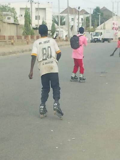 Three people are rollerblading down a street. One wears a white shirt with a yellow collar, "M. FELE" on the top, and "80 AZ WARRIORS" below. The person wears dark pants. Another rollerblader wears pink with a black backpack and red pants. A third person skates in the distance. Buildings are visible behind the skaters. A white truck is parked near the background.
