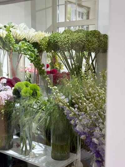 The image shows a display of flowers in glass vases, arranged on a white shelf. The flowers are diverse in type and color, with prominent white, green, and purple blooms. In the background, a reflection in the glass shows a building with windows, resembling a storefront. The overall scene suggests a flower shop or a floral arrangement display. There are no people visible in the image. The location is specified as Bishkek City, Kyrgyzstan.