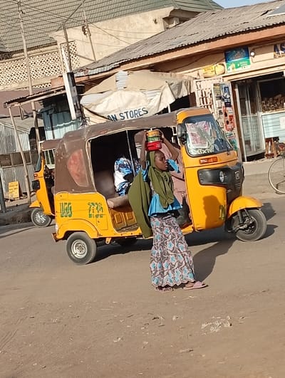 The image depicts a street scene in Gwagwarwa, Nigeria, under direct sunlight, evidenced by shadows cast to the right. A dark-skinned woman walks on a dusty asphalt road, balancing a stack of three lidded containers (red, orange, green) on her head. She wears an olive-green top over a blue garment, a long skirt with a blue, grey, and white floral pattern, and pink sandals. Adjacent to her on the left is a yellow three-wheeled auto-rickshaw, equipped with a brown fabric canopy and displaying "UGG" on its side. A person in a light-colored shirt is visible in the driver's seat of this vehicle. Further to the left and behind, a second yellow three-wheeled auto-rickshaw is partially visible, with a passenger in a blue and white patterned garment seated inside. The background consists of several commercial buildings featuring corrugated metal roofs. One structure includes a beige tarp awning with a partial "AN STORES" label, while another presents a shopfront with a glass door displaying merchandise.