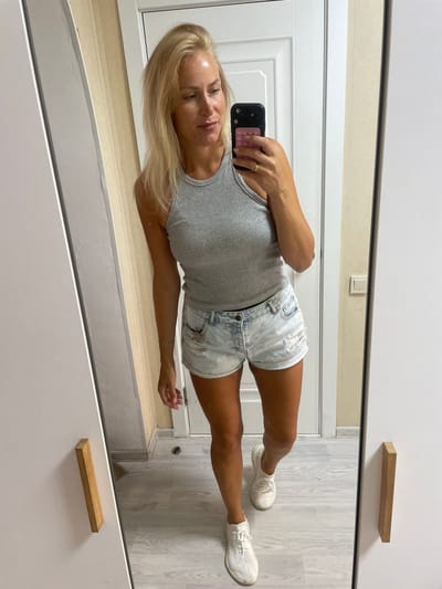 A full-body mirror selfie shows a blonde woman in a grey ribbed tank top and light-wash denim shorts with rips. She is wearing white athletic shoes. The woman is standing in what appears to be an apartment, with a white door visible in the background. The room has light wood-toned flooring and light-colored walls. The woman's pose and the setting suggest a casual, everyday moment. There is no visual information that would allow for determining a specific location within the city of Baranovka, Russia.