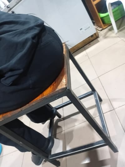 The image shows a person sitting on a wooden and metal stool in what appears to be an indoor setting with tiled flooring. The person is wearing dark clothing and black shoes. In the background, a white appliance, possibly a freezer, and a wooden shelf with a green basket are visible. The lighting suggests it could be daytime. The scene is located in Howayi, Nigeria.