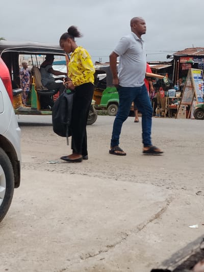 The image depicts a street scene in what appears to be the city of Aba, Nigeria.
A woman in a yellow floral shirt and black pants stands on the left side of the frame, carrying a black bag. Next to her on the right, a man in a gray polo shirt and blue jeans is walking. They are both on a paved road.
Behind them, a green and yellow auto-rickshaw is visible, with at least two passengers inside. A market stall with signage and various items for sale is visible in the background. Other people and vehicles are present in the scene, suggesting a busy commercial area.
The sky is overcast, and the lighting is diffused. The foreground shows the pavement's details, including cracks and imperfections. A portion of a vehicle's tire is visible on the bottom left.