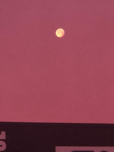 The image shows a pale orange circle, presumably the moon, against a muted pink sky. In the lower portion of the frame, a dark, horizontal band is visible, with faint white letters on it. This band appears to be a sign or billboard. The environment appears to be outdoors. No specific landmarks of Leudelange, Luxembourg are identifiable.