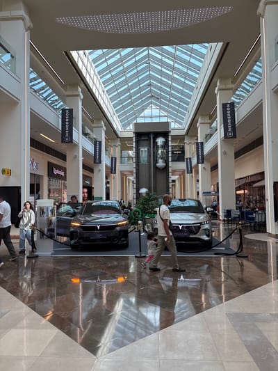 The image depicts a modern indoor shopping mall, likely in Yerevan, Armenia, given the presence of signage in Armenian and the architectural style. The scene is dominated by a spacious atrium with a glass-paneled roof, allowing natural light to flood the area. Two cars, possibly electric vehicles, are prominently displayed on a raised platform in the center of the mall. One car is black with its doors open, while the other is silver. Both vehicles have illuminated grilles and distinctive logos.

Several individuals are visible in the image. In the foreground, a man with a bald head and a beard is walking from left to right, holding a small child's hand. The child is wearing a pink outfit. To the left, a woman with curly dark hair is talking on her phone. Other shoppers are scattered throughout the mall, some browsing stores, others seated at tables in a café visible in the background to the right. The mall's interior features polished marble floors that reflect the ambient light and architectural elements. Numerous columns, some adorned with black banners featuring the "H HENDERSON" logo, support the upper levels of the mall. Storefronts line the walls, with one displaying "MAC" cosmetics. An elevator with a glass shaft is also visible on the left side of the atrium. The overall impression is one of a clean, well-lit, and contemporary commercial space.