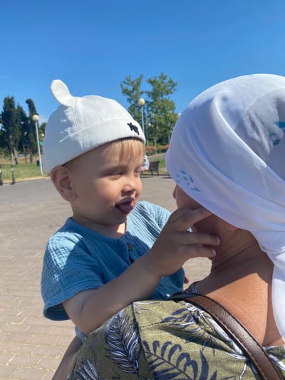 An infant boy wearing a white hat with a rabbit ear-like protrusion and a light blue textured short-sleeved shirt is held by an adult. The infant's tongue is sticking out. The adult is wearing a white headscarf with a subtle blue floral pattern and a green patterned top. The scene takes place outdoors on a sunny day with a paved walkway and trees in the background. The overall atmosphere is casual and affectionate. The image is taken at a close-up perspective, focusing on the interaction between the infant and the adult.