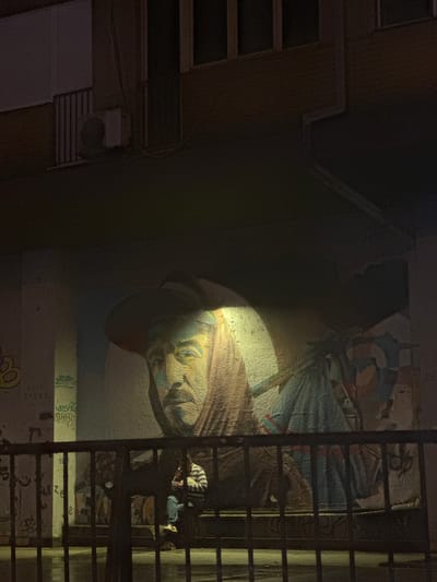 A person sits on a bench in front of a mural in Belgrade, Serbia, at night. The mural depicts a large face wearing a hat and a hooded garment, illuminated by a spotlight. The person is wearing a striped shirt and blue jeans, and is looking down at something in their hands. The scene is framed by a black metal fence. The wall also features other graffiti tags. An air conditioning unit is visible on the building above. The ground appears wet, suggesting it may have recently rained.