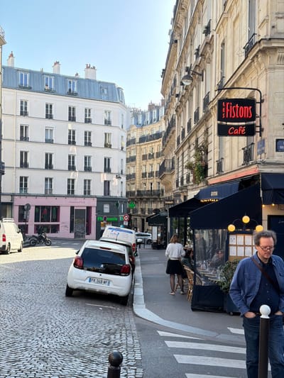 An outdoor daylight scene depicts a cobblestone street intersection in Paris, France, specifically at the corner of Rue d'Argenteuil and Rue Saint-Honoré in the 1st arrondissement. The sky is clear and blue, and sunlight illuminates the scene.
On the right side of the image, a tall, light-colored Haussmannian building features multiple stories with windows and decorative ironwork balconies. At its ground floor corner, the "Les Fistons Café" is prominent, identified by a retro-style black neon sign displaying "LES FISTONS" in red script above "Café" in red capital letters. The café has a dark blue awning extending over an outdoor seating area. This area is furnished with small tables and bamboo-colored chairs, partially enclosed by a black barrier with transparent plastic panels. Spherical yellow lamps are suspended from the awning's structure. Patrons are seated at the tables, some partially visible.
In the mid-ground, a woman with light hair, wearing a white sweater and black skirt, stands near the café's outdoor seating, facing away from the viewer. In the immediate foreground, to the right, a man with wavy brown hair and glasses, wearing a blue button-down shirt and blue jeans, walks leftwards with his hands in his pockets, a dark strap crossing his chest.
On the left side of the image, across the cobblestone street, stands a multi-story white building with numerous windows and a blue-grey mansard roof. Its ground floor features a pink storefront labeled "Antoine & Lili," displaying merchandise through a window. Adjacent to it, a green "Pharmacie" sign is visible.
The cobblestone street features a white crosswalk. Several vehicles are parked on the left side of the street, partially on the curb. A white Peugeot 108 with French license plate "FK-348-WF" is most prominent, parked facing away from the viewer. Behind it, a white delivery van is parked. Further down the street, a parked motorcycle or scooter is visible. A black bollard with a white top is present on the sidewalk in the bottom right corner. Other Haussmannian buildings extend into the background.
