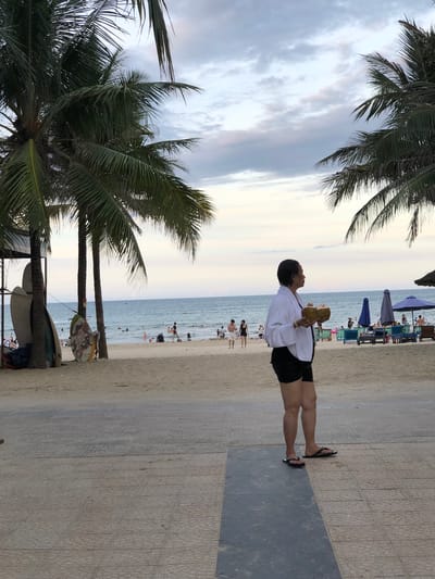 The image depicts a casual scene on a beach in Da Nang, Vietnam. The primary subject is a person standing on a paved walkway, holding two coconuts. They are wearing a white, possibly terrycloth, shirt and black shorts, and black flip-flops. The person is looking upwards and to their left, with their head tilted back.
Behind the person, the beach stretches out towards the ocean. The sand is a light beige color, and the water appears calm and blue, with gentle waves. Several other people are visible on the beach, some wading in the water, others sitting on beach chairs or walking along the shore. Beach umbrellas, in shades of blue and purple, are dotted across the sand, indicating a typical beach resort setting.
In the foreground, on the left side of the image, are the fronds of a palm tree, with its leaves dark green against a cloudy sky. The sky itself is overcast with soft, diffused light, suggesting it is either early morning or late afternoon. The paved walkway where the person stands is made of textured concrete tiles. The overall mood is relaxed and summery. There is no visible text in the image.