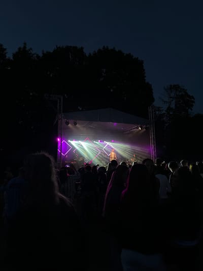 The media file depicts an outdoor concert at night. The foreground is dark, with the silhouettes of a crowd of people visible. The crowd is facing a brightly lit stage where a band is performing. The stage is adorned with white and purple lights that create beams of light extending outwards. A performer, possibly a singer, is visible on stage, illuminated by the stage lights. The backdrop is a dark sky, with the silhouette of trees on the left side of the frame. The overall atmosphere is energetic and lively, typical of a concert setting. The time of day is clearly night, and the weather appears clear. The location is Panevėžys, Lithuania. No visible text is present in the image.