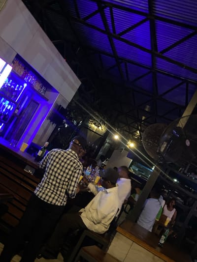 The image shows a bar in Dar es Salaam, Tanzania, located at the coordinates -6.727 latitude and 39.142 longitude. The bar is lit by a blue neon light, which reflects on the ceiling of the bar. There are several people in the bar, including a man wearing a black and white plaid shirt and a man wearing a white shirt. Both men are holding yellow cups, possibly containing a drink. There is a long wooden bar to the left of the image and shelves on the right side. There is a large fan in the corner of the bar and many more people are visible in the background. The bar has a modern feel and the environment is relaxed and casual.