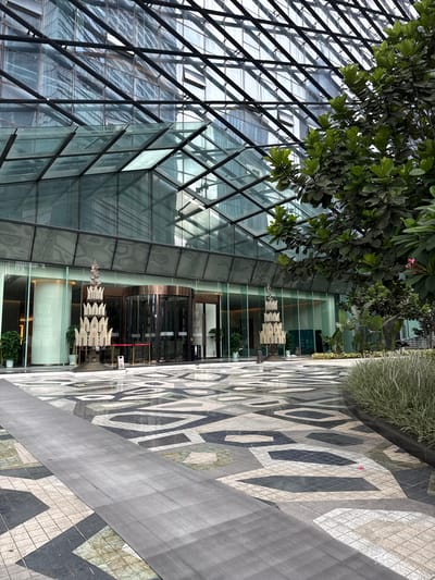 The image depicts a modern architectural building with a glass facade and a geometric patterned outdoor pavement. Green foliage from trees is visible in the upper left portion of the frame, casting shadows on the glass structure. The building's exterior is characterized by a lattice of black metal beams supporting large glass panels, creating a reflective and somewhat abstract surface.

In the foreground, the pavement features a repeating pattern of interconnected geometric shapes in shades of gray, black, and white. A dark gray walkway runs diagonally across this patterned area. Beyond the glass facade, a revolving door entrance to the building is visible. Inside, the interior appears to be a well-lit lobby or atrium, with modern lighting fixtures and some potted plants. A piece of modern art, consisting of stacked, layered elements, is suspended in front of the entrance.

The scene appears to be an outdoor space in a contemporary urban environment, likely during daylight hours given the lighting. The overall impression is one of sleek, modern design and a focus on natural light and geometric aesthetics. There are no people clearly visible in the frame, suggesting an unoccupied moment or a focus on the architecture and design elements. There is no visible text in the image.