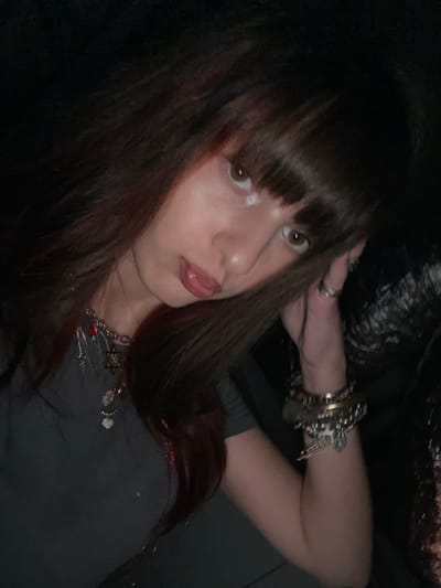 A young woman with dark, shoulder-length hair and blunt bangs poses indoors. She wears multiple bracelets and a necklace with charms, including a Star of David. She glances towards the camera, with her head tilted slightly. The background is dark and indistinct, but appears to be a textured, possibly metallic, surface. Her expression is neutral, with slightly parted lips.