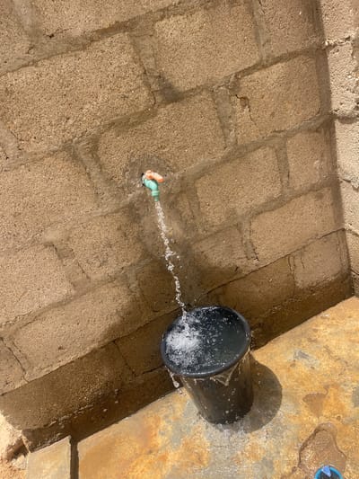 The image depicts an outdoor water collection point, possibly in Unguwar Gabas, Nigeria. The scene features a rough, concrete brick-paved area. A green tap is affixed to the wall, and water flows from it into a black plastic bucket. The water spills over the rim of the bucket, wetting the surrounding concrete and draining into the crevice by the wall. The area appears utilitarian and likely serves as a water source for the local community.